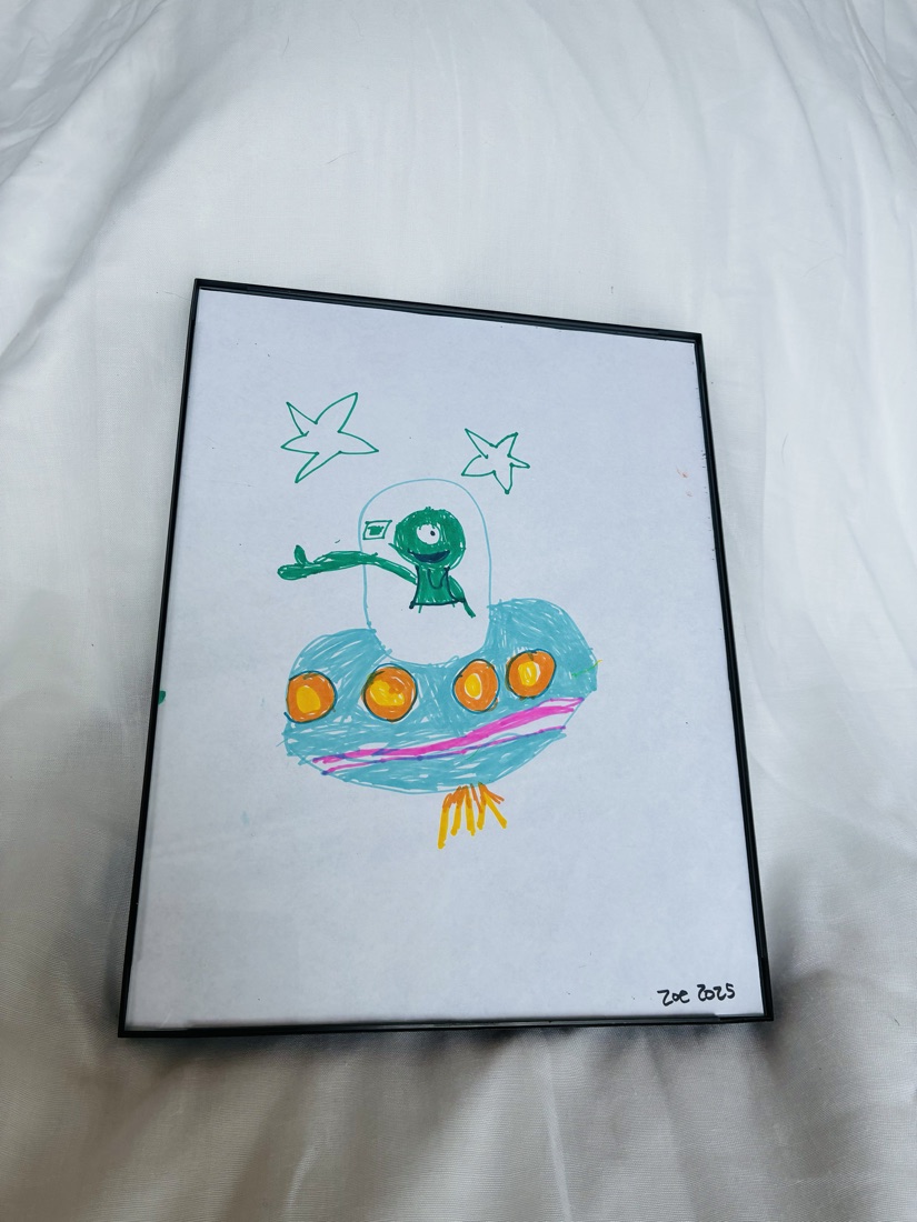 Marker drawing of a green one-eyed alien waving from a blue flying saucer with orange lights, under two green stars