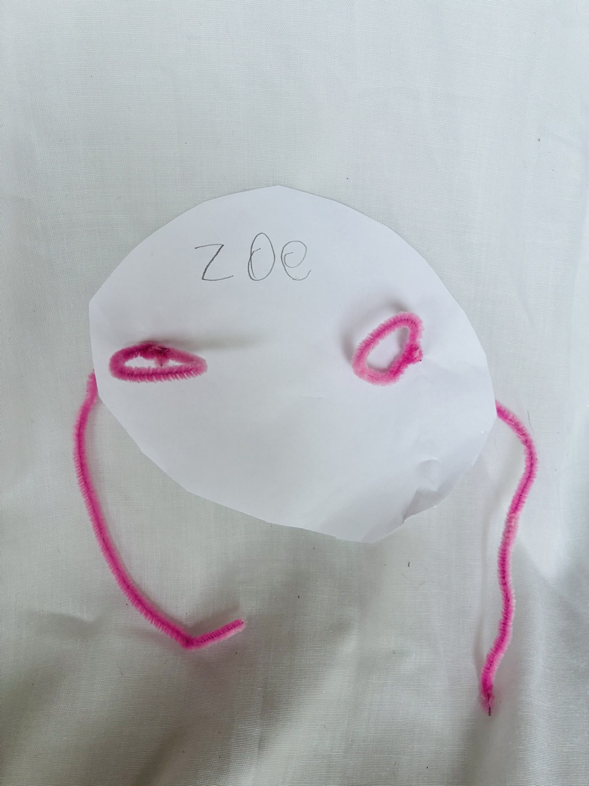 A paper mask with eye and mouth holes outlined in pink pipe cleaners, with pink ties, labeled Zoe
