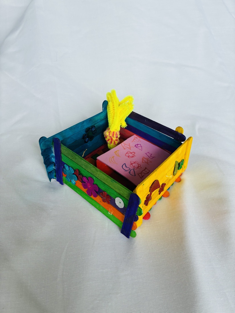A small handmade box made of painted popsicle sticks decorated with felt flowers, holding pink paper inside